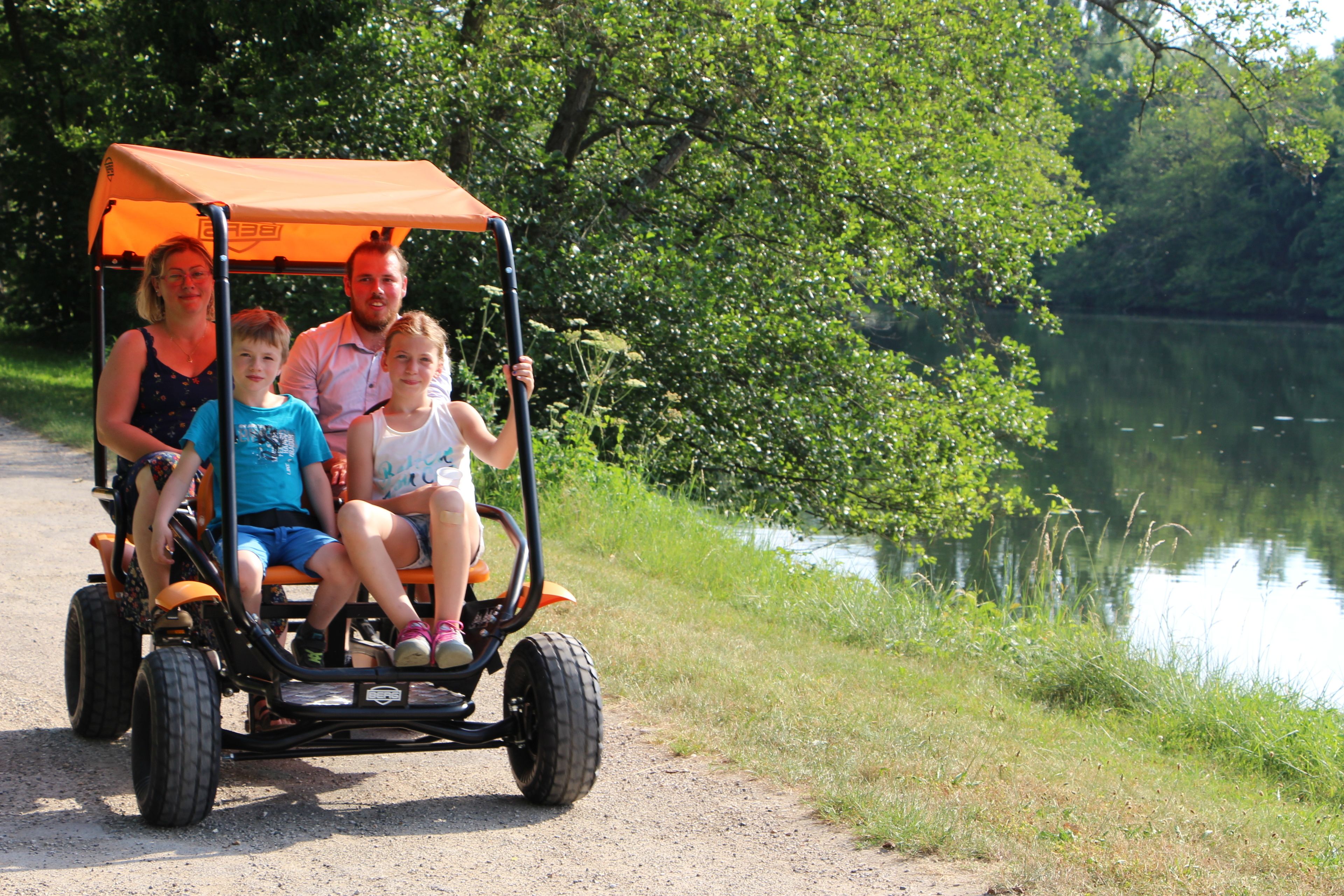 A family of four on a small, open-top vehicle with a canopy, traveling along a path beside a body of water, surrounded by greenery.