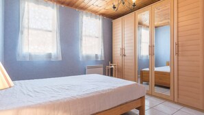 A bedroom with a wooden bed, two windows with sheer curtains, a wardrobe with a mirror, and a tiled floor.