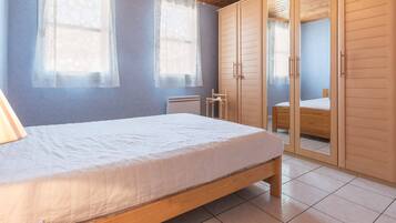 A bedroom with a wooden bed, two windows with sheer curtains, a wardrobe with a mirror, and a tiled floor.