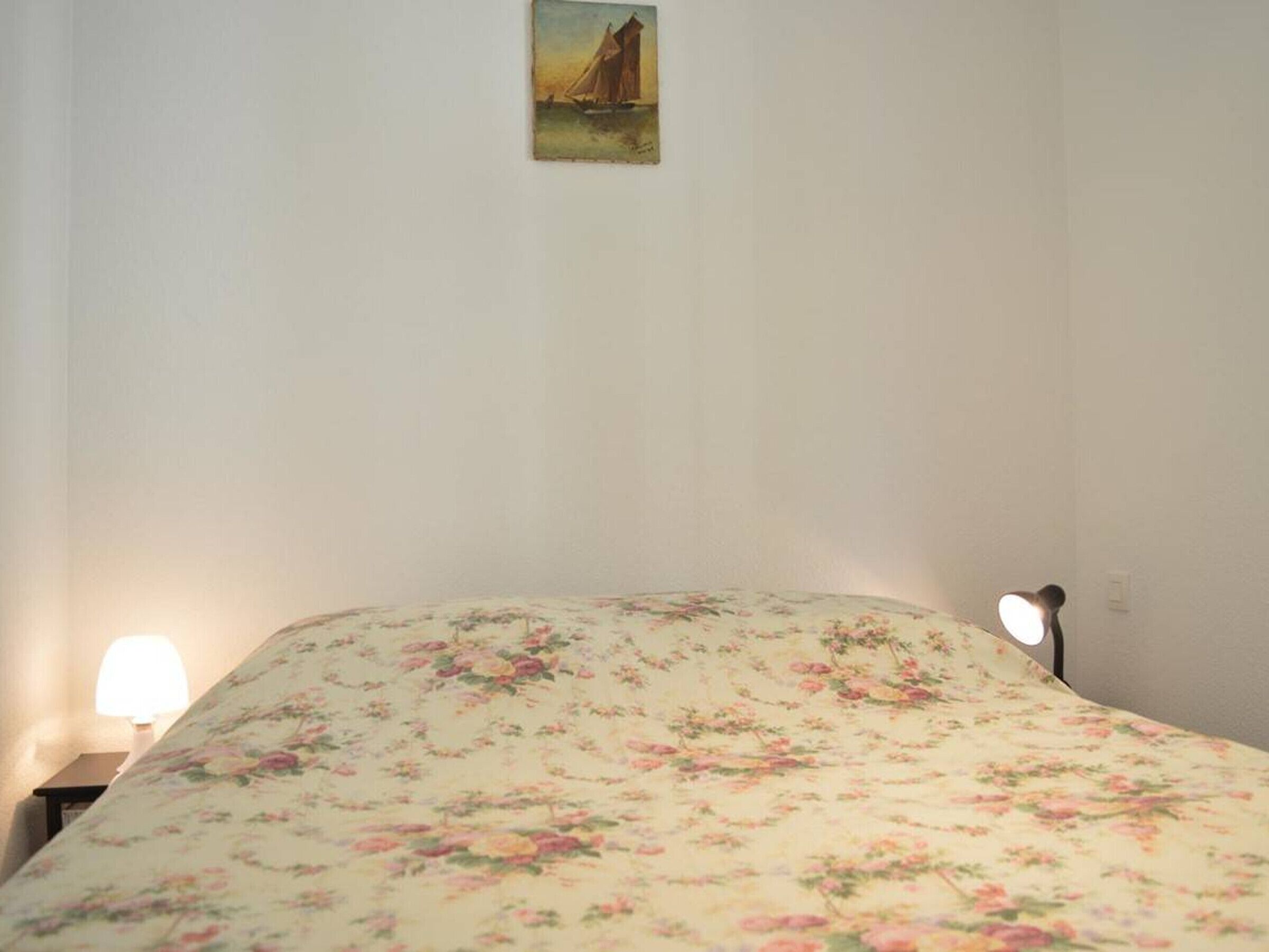 A bedroom with a floral bedspread, a nightstand with a lamp, and a small painting on the wall.