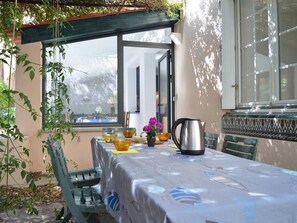 A covered outdoor dining area with a table set for two, a metal pitcher, and a vase with flowers.
