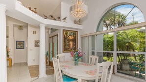 A dining area with a round table, white chairs, and a chandelier. There is a large window with a view of greenery outside. - Pet Friendly Coastal Retreat | Canal Access | Beach Nearby (New Smyrna Beach)