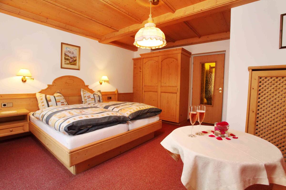 Une chambre à coucher avec un cadre en bois, comprenant un lit, des lampes de chevet, une armoire en bois et une table sur laquelle se trouvent une coupe de champagne et des pétales de rose.