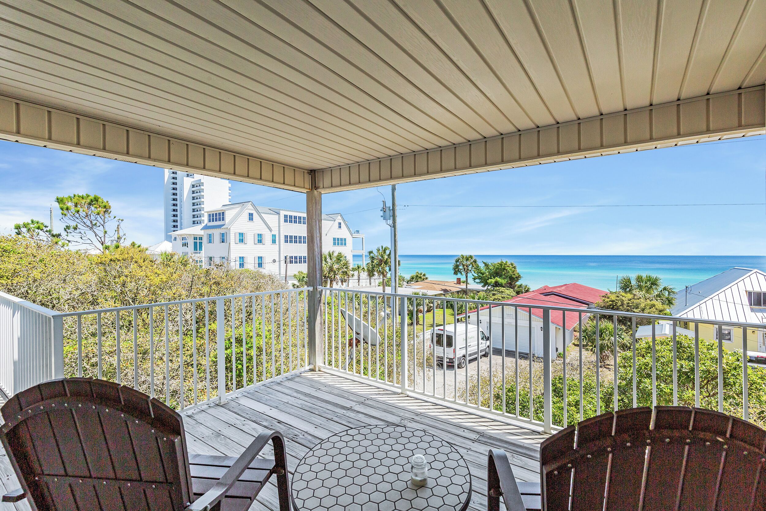 A covered balcony with a view of the ocean, featuring two Adirondack chairs and a hexagonal table.