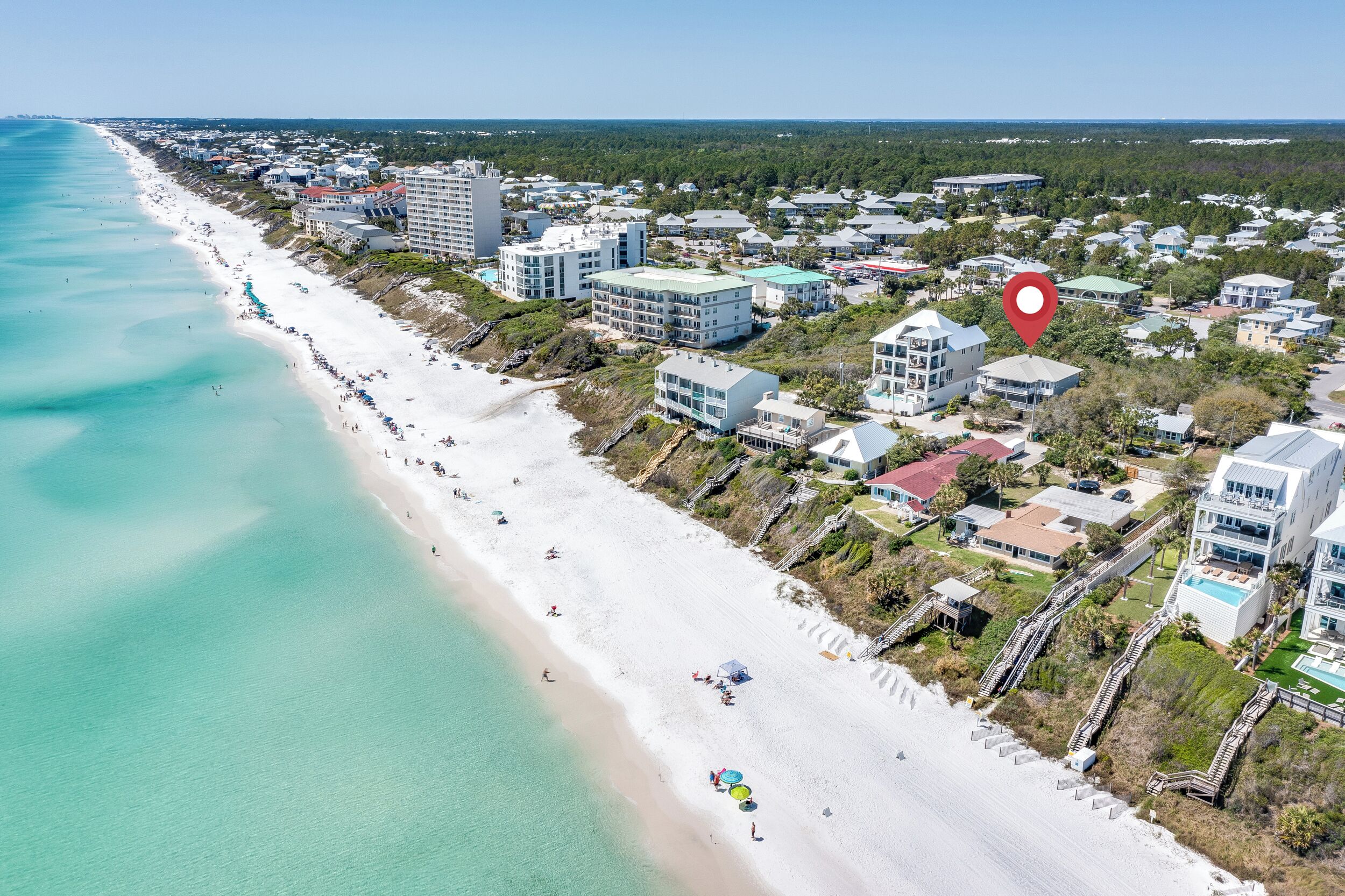 An aerial view of a coastal area with a long sandy beach, beachfront properties, and a red location marker.