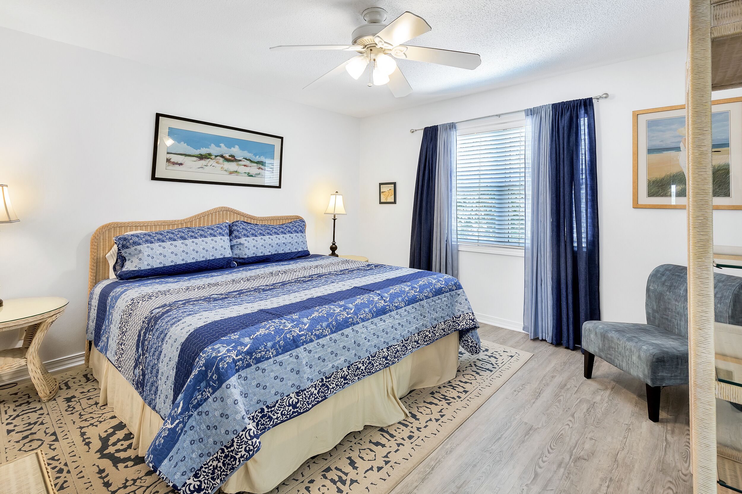 A bedroom with a bed, a chair, a nightstand, a lamp, a ceiling fan, and a window with curtains.