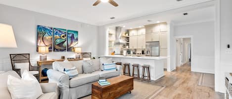 A modern living room with a sofa, coffee table, and artwork. A kitchen with white cabinets and a bar area is visible in the background.