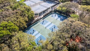 Ein Tennisplatz im Freien, umgeben von dichtem Baumbestand, mit einem Pool und einem Gebäude im Hintergrund.