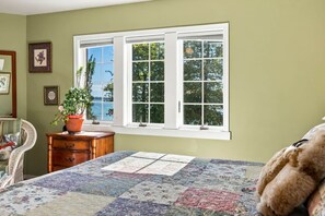 A bedroom with a large window, a bed with a quilt, a dresser, a chair, and a potted plant.
