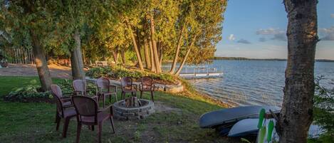 Ein Sitzbereich am Seeufer mit Stühlen und einem Feuerkorb, umgeben von Bäumen und mit Blick auf das Wasser.