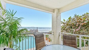 Ein Balkon mit Glastisch und -stühlen, der einen Blick auf einen Pool und einen Strand mit einer Brücke in der Ferne bietet.