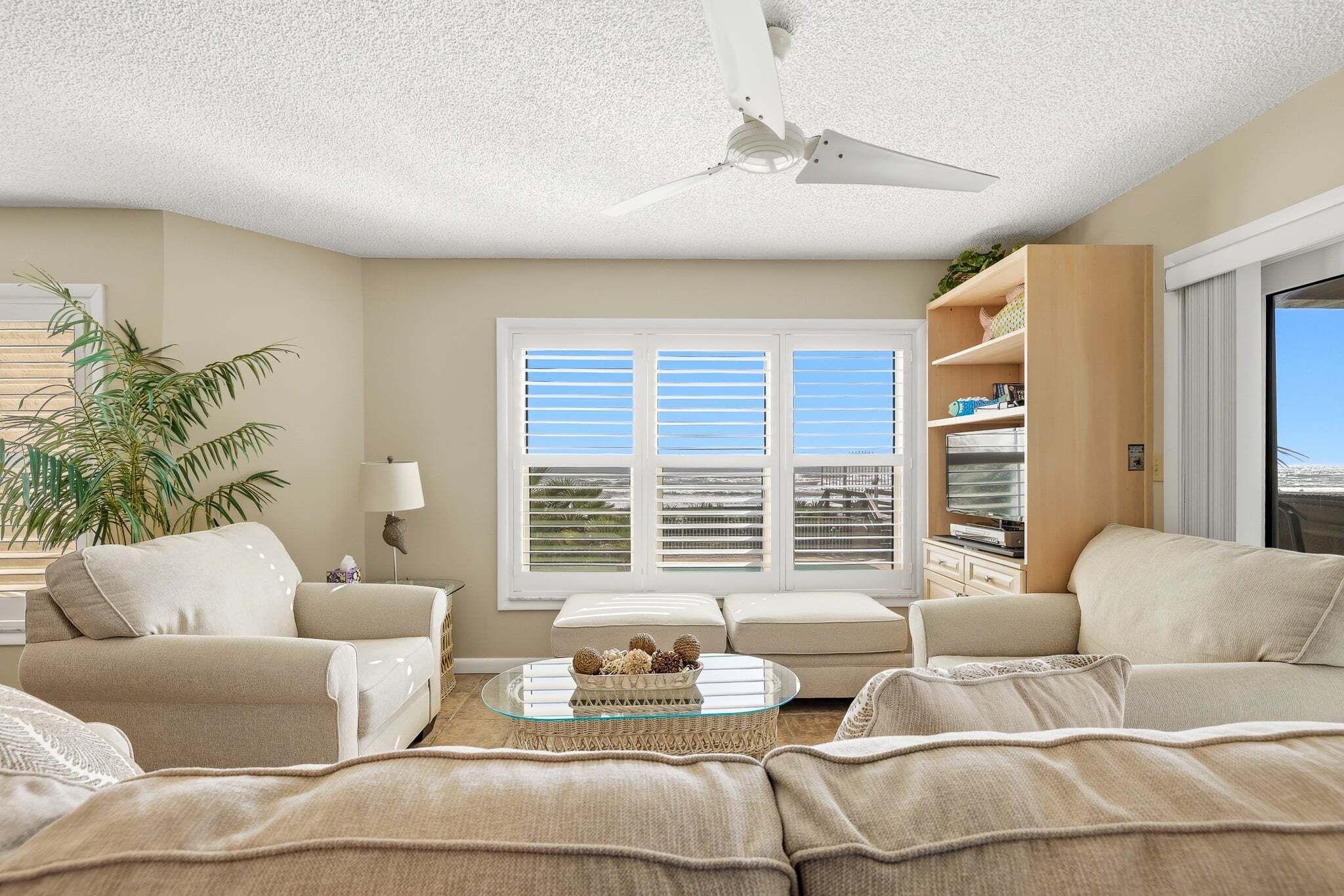 Un salon avec un ventilateur de plafond, une grande fenêtre équipée de stores, un canapé, une table basse et une bibliothèque.