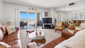 A living room with a glass coffee table, leather sofas, a flat-screen TV, and a balcony with a view.
