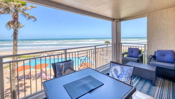 A balcony with a view of the beach, outdoor seating, and a pool.