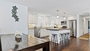 Une cuisine moderne avec des meubles blancs, des appareils en acier inoxydable et une table à manger en bois, accompagnée de tabourets de bar.