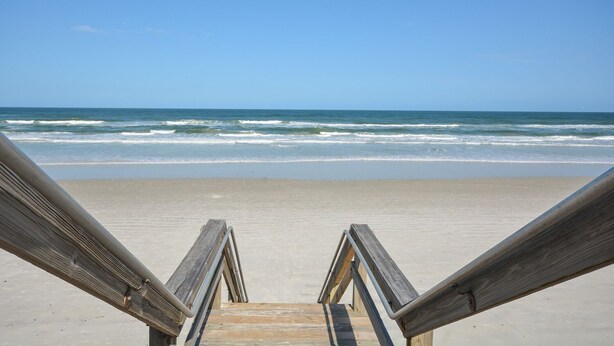 Une promenade en bois mène à une plage de sable, avec l’océan et ses vagues en arrière-plan.