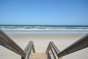 Une promenade en bois mène à une plage de sable, avec l’océan et ses vagues en arrière-plan.
