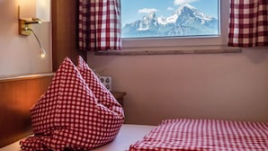 Une chambre d’hôtel avec un lit, des rideaux à carreaux, une fenêtre donnant sur la montagne et une lampe fixée au mur.
