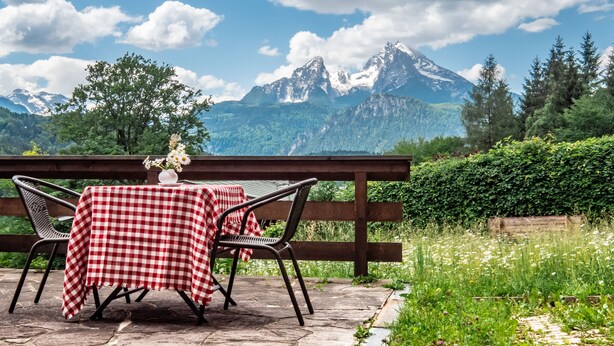 Une table recouverte d’une nappe à carreaux rouges et blancs, deux chaises et un vase rempli de fleurs, le tout placé devant un paysage de montagne avec une clôture en bois.