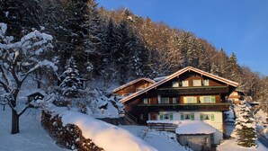 Un chalet en bois à deux étages, doté d’un balcon, entouré d’arbres couverts de neige et surplombant un ciel dégagé.