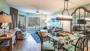 A dining area with a glass table, chairs, and a chandelier. There is a ceiling fan, a window with blinds, and a painting on the wall. - $150 activities - E-gift card when you book - Lakefront Screened in Porch | KLC 112 (Myrtle Beach)