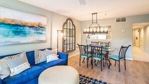 A modern living room with a blue sofa, a dining table with chairs, and a kitchen in the background. - $150 activities - E-gift card when you book - Lakefront Screened in Porch | KLC 112 (Myrtle Beach)