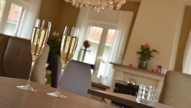 A dining area with two champagne flutes on a table, a chandelier, and a fireplace in the background. - Amylia (De Haan)
