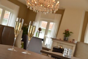 A dining area with two champagne flutes on a table, a chandelier, and a fireplace in the background. - Amylia (De Haan)