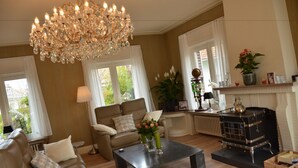 A living room with a chandelier, a sofa, a coffee table, a fireplace, and a window with curtains. - Amylia (De Haan)
