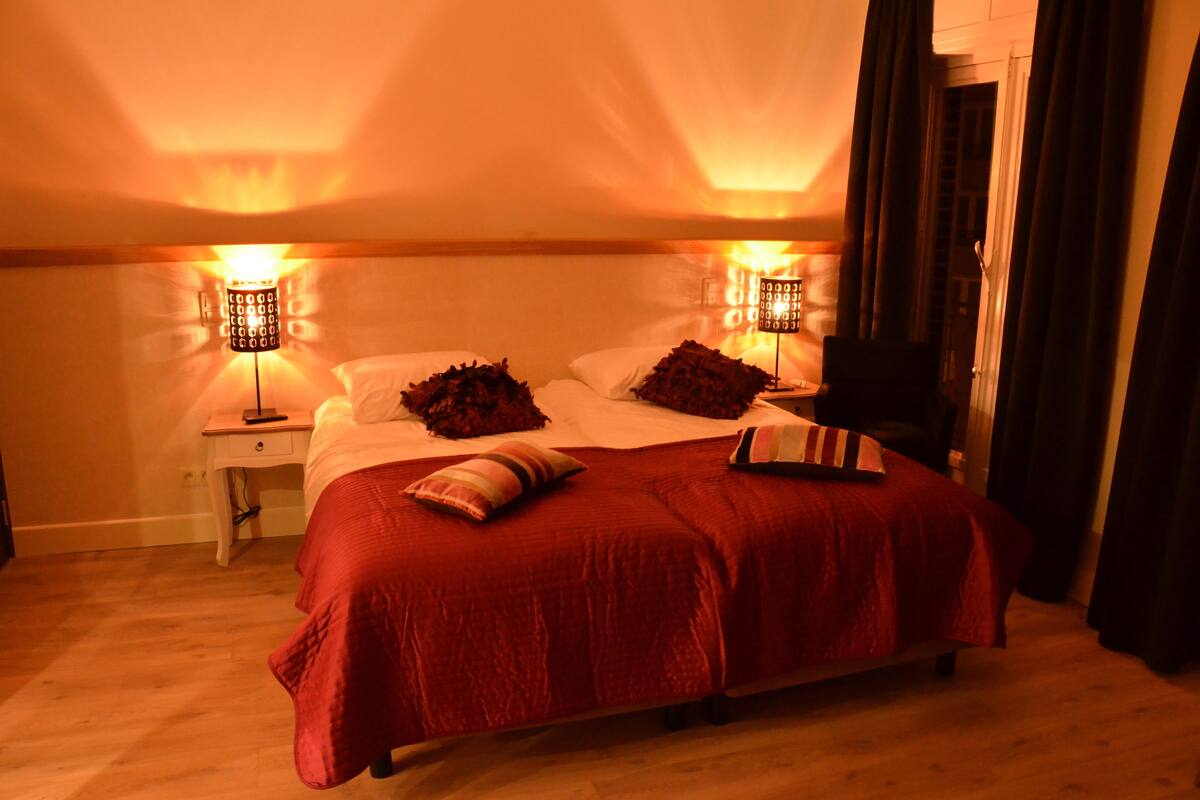 A hotel room with a large bed, two bedside lamps, a nightstand, and a view of the sea through the window.