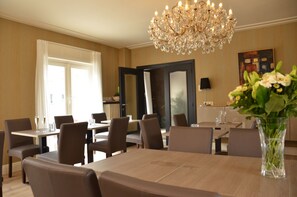 A dining area with a chandelier, tables, chairs, and a vase with flowers. - Amylia (De Haan)