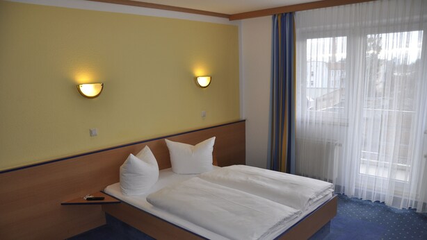A hotel room with a bed, two wall-mounted lights, a window with sheer curtains, and a view of buildings outside. - Sleep & Go Hotel Magdeburg (Magdeburg)