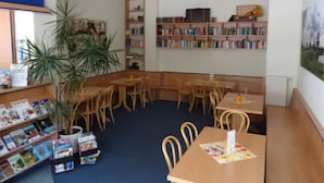 A library with bookshelves, tables, chairs, and a potted plant. - Sleep & Go Hotel Magdeburg (Magdeburg)