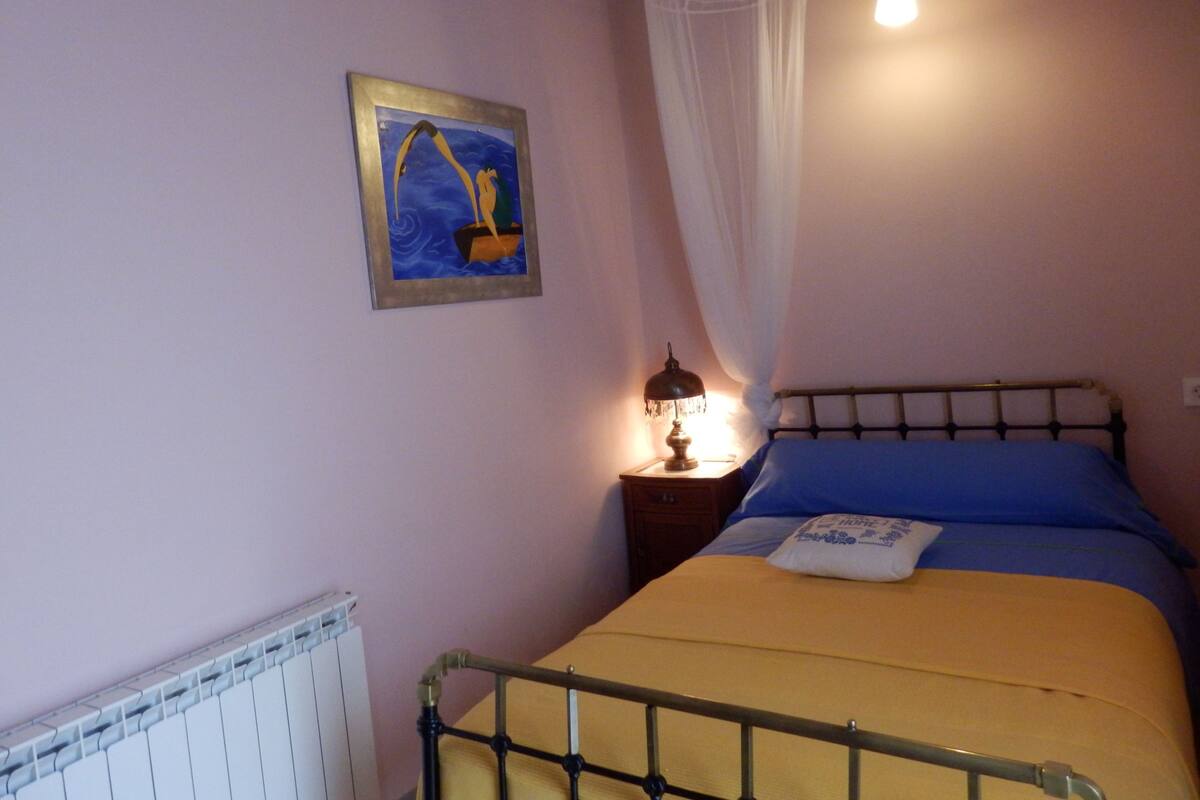 A bedroom with a bed, a nightstand, a lamp, a picture on the wall, and a window with curtains.