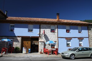 A two-story building with a stone facade and a tiled roof. There is a car parked in front, and outdoor seating with red chairs and blue umbrellas. - CasaCándana (La Vecilla)