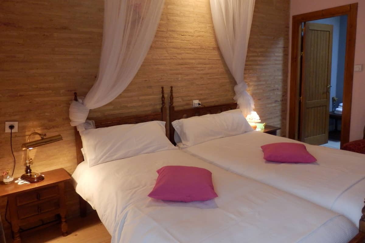 A double bed with white linens and two pink pillows, a bedside table with a lamp, and a wooden headboard.