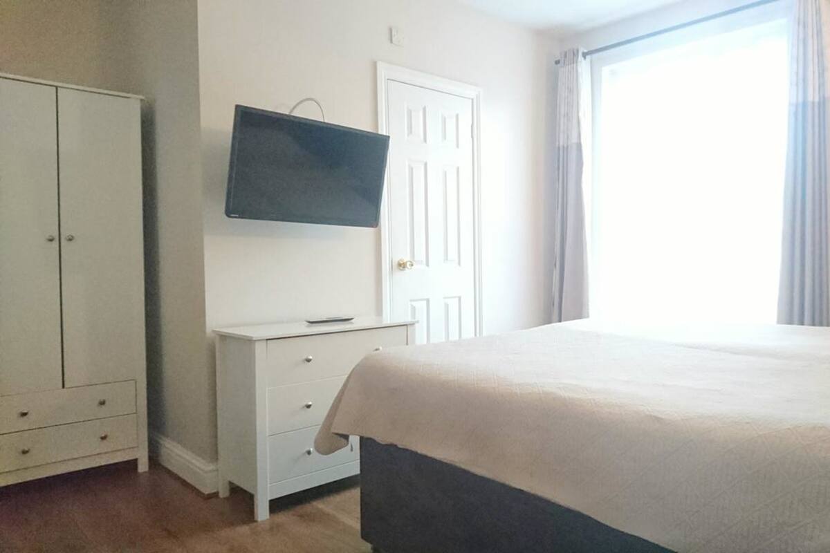 A bedroom with a bed, a TV mounted on the wall, a wardrobe, and a window with curtains.