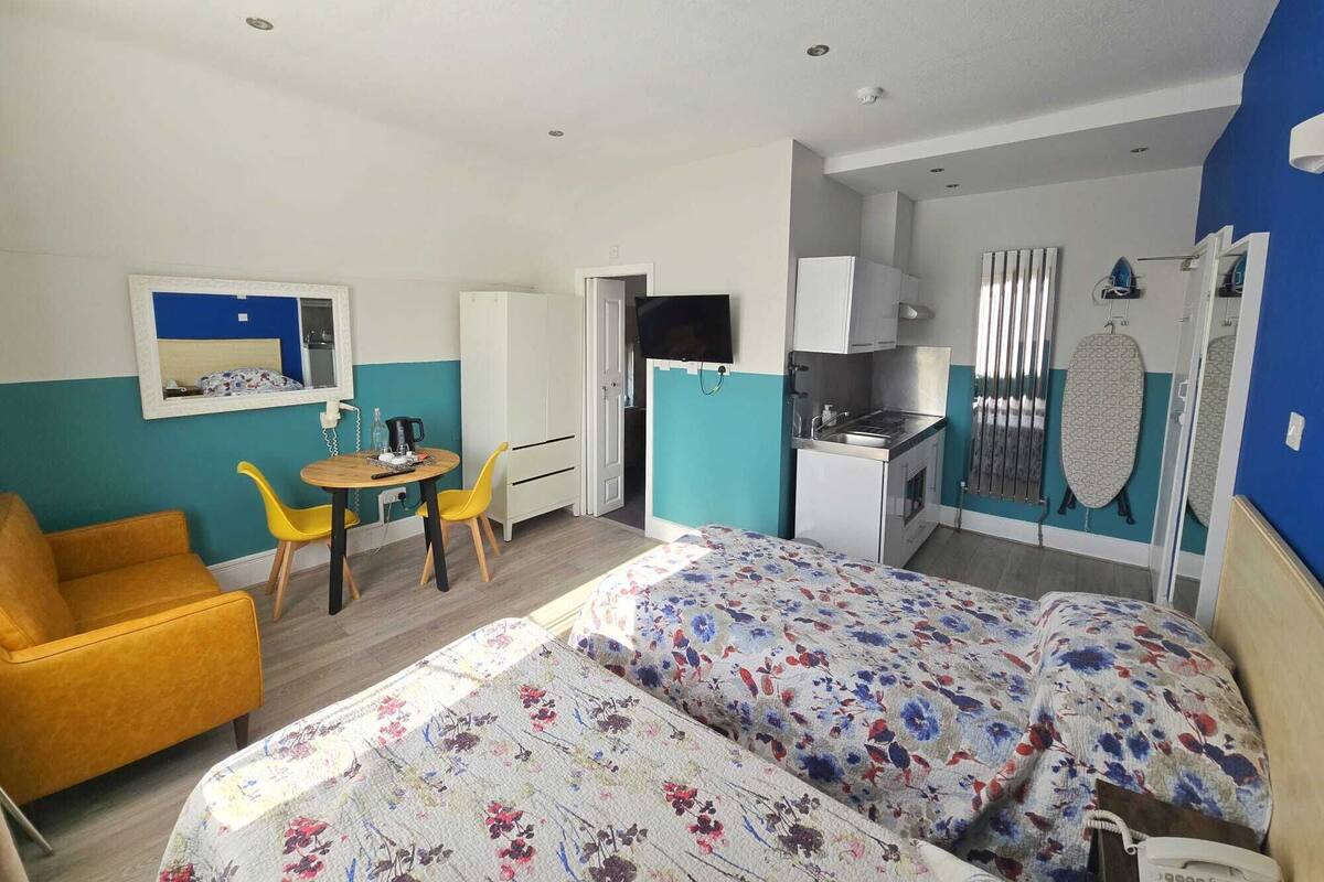 A compact room with a bed, a small dining table, a yellow chair, a kitchenette, and a TV.