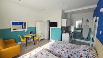 A compact room with a bed, a small dining table, a yellow chair, a kitchenette, and a TV.