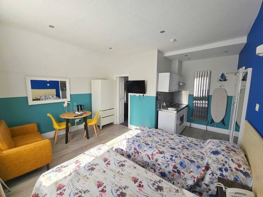A compact room with a bed, a small dining table, a yellow chair, a kitchenette, and a TV.
