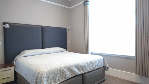 Una cama bien hecha en una habitación de hotel con cabecera, mesita de noche y ventana con cortinas.