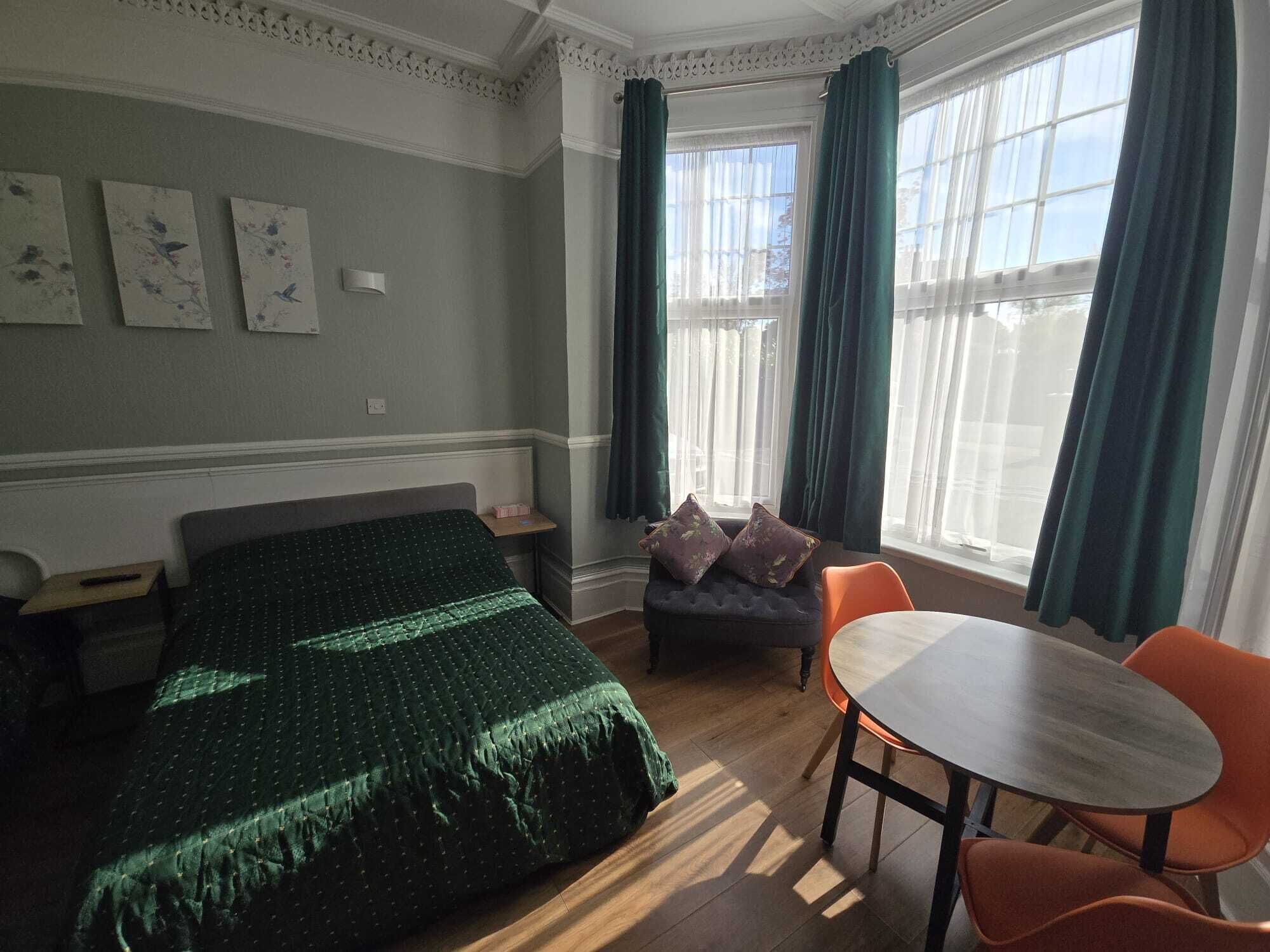 Habitación de hotel con cama, una mesita, dos sillas, una mesita de noche y una ventana con cortinas.