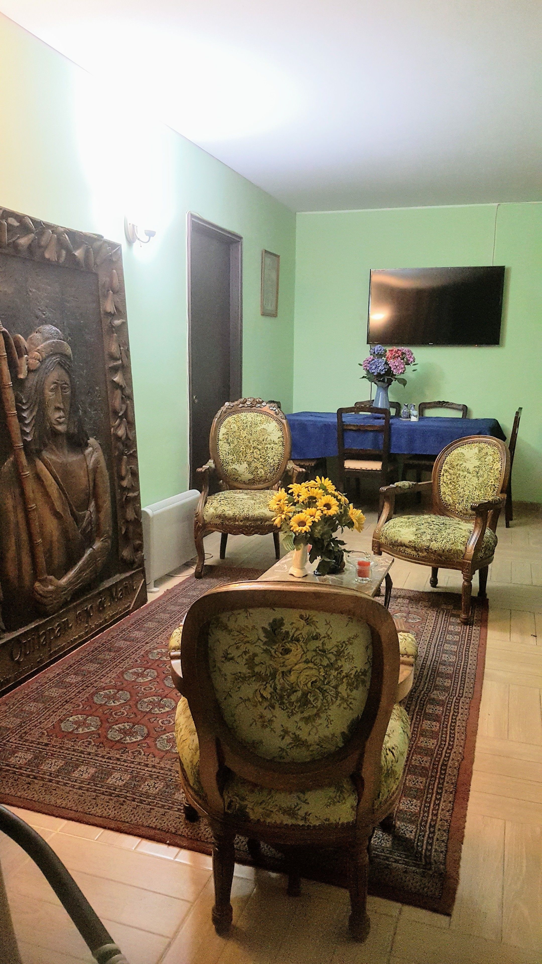 A living room with two ornate chairs, a coffee table with a vase of sunflowers, a large Buddha statue, a dining table with chairs, and a flat-screen TV on the wall.