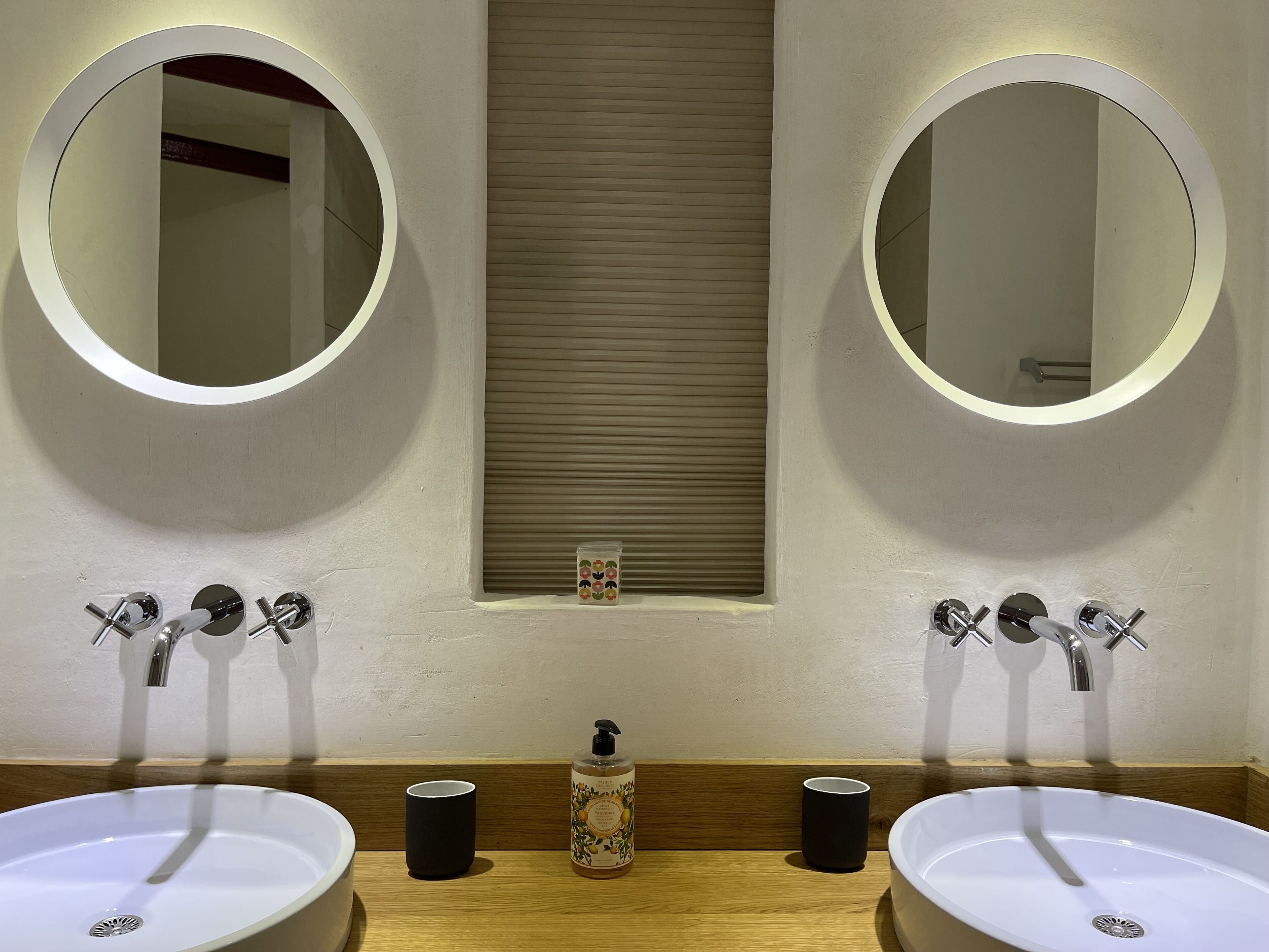 Un baño con dos lavamanos, dos espejos redondos y una ventana con persianas.
