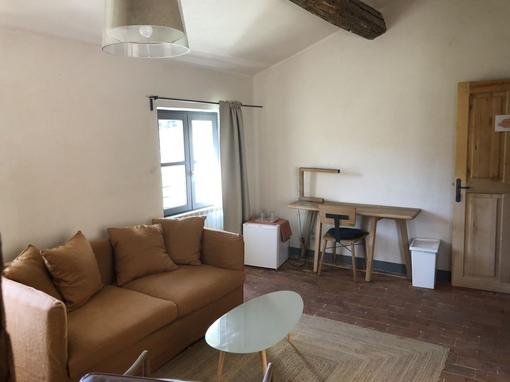 Una sala de estar con un sofá marrón, una mesa de centro de vidrio, un escritorio con una silla y una ventana con cortinas.