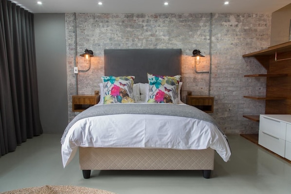 Une chambre à coucher moderne avec un grand lit, deux tables de chevet, une commode en bois et un mur en briques.