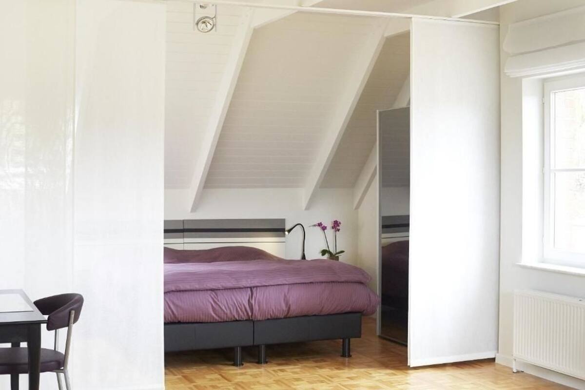A bedroom with a sloped ceiling, a bed with a purple bedspread, a small table with a chair, and a window with blinds.