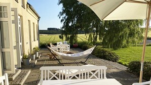 Un patio al aire libre con mobiliario blanco, incluyendo una mesa y sillas, bajo un gran paraguas.