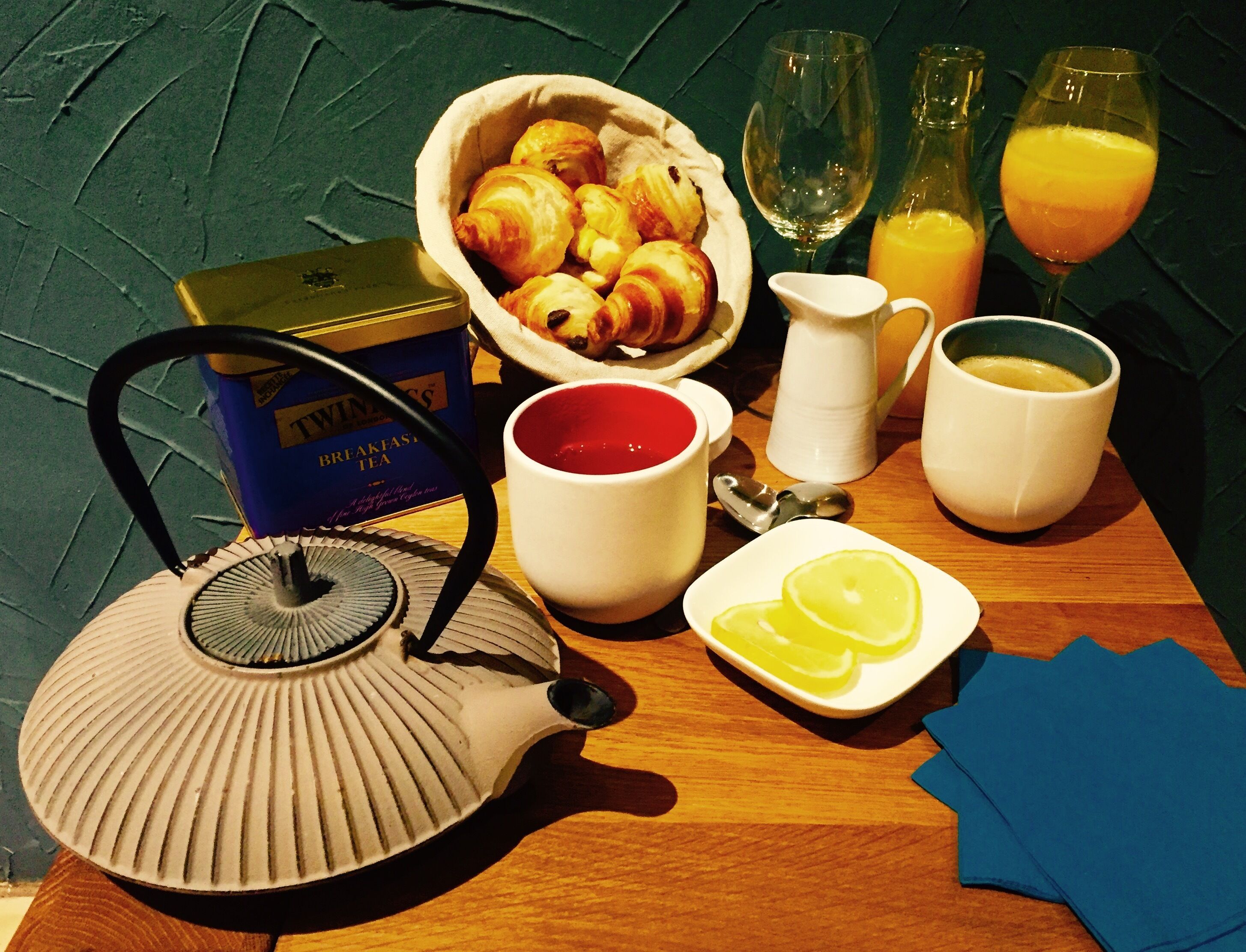 Una mesa de desayuno con cruasanes, una tetera, tazas y vasos de jugo de naranja.