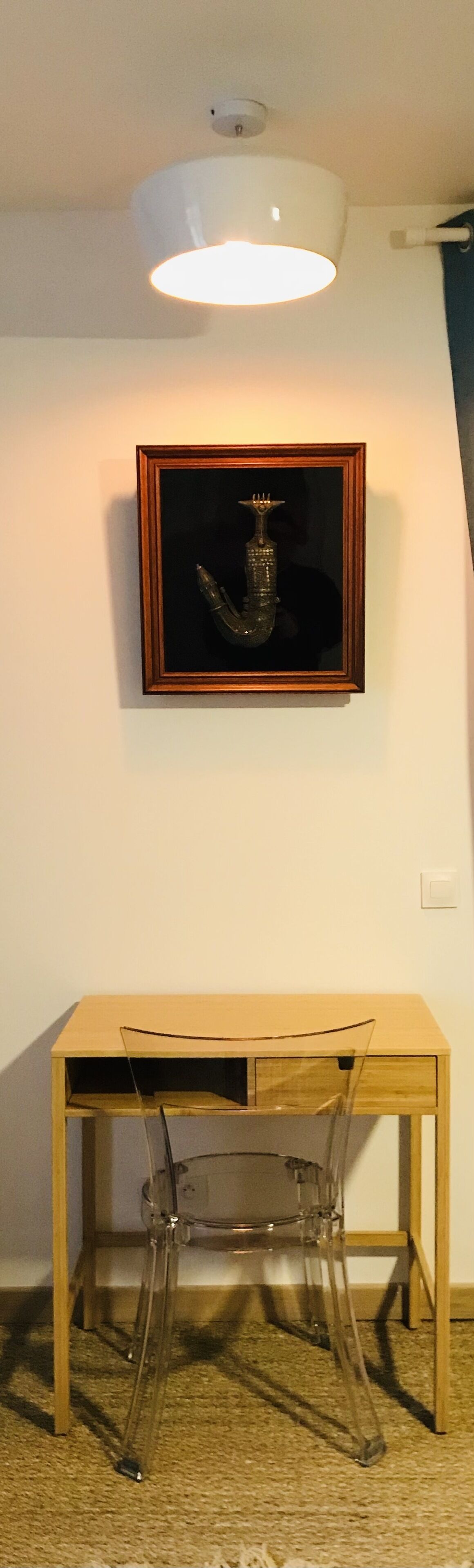 Une pièce avec un bureau en bois, une chaise transparente, un objet encadré accroché au mur et un luminaire au plafond.